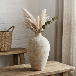 May include: A textured, off-white vase with a wide base and narrow neck, filled with fluffy, beige pampas grass and a sprig of green eucalyptus. The vase sits on a rustic wooden table. The vase is a decorative item.
