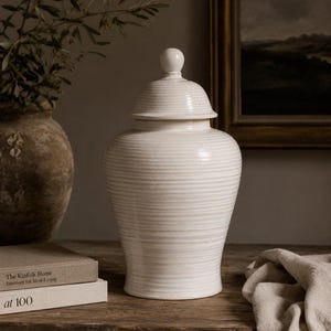 May include: A cream-coloured ceramic jar with a lid, showing horizontal ribbed detailing. The jar is on a wooden surface, with a neutral background. A decorative item for home interiors.