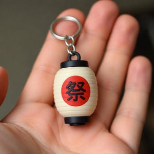 Pode incluir: Um chaveiro com uma lanterna japonesa em miniatura. A lanterna é creme com um círculo vermelho contendo caracteres japoneses pretos. A parte superior e inferior são pretas e pendura de um chaveiro prateado.