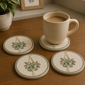 May include: Three round off-white coasters with a sage green border and a raised relief design of a potted plant in a macrame hanger. One coaster supports a matching off-white ceramic mug filled with a light brown beverage, while two others sit nearby.