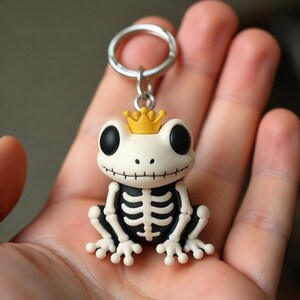 May include: A keychain featuring a white frog with a black skeleton print on its body, large black eyes, and a stitched mouth. The frog wears a small yellow crown on its head and is attached to a circular silver metal ring for easy attachment.