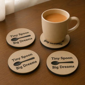 Può includere: Tre sottobicchieri rotondi color beige con bordi neri, ognuno caratterizzato da un grafico a cucchiaio nero e dal testo "Tiny Spoon Big Dreams" in caratteri neri. Una tazza in ceramica beige riempita con una bevanda marrone chiaro poggia su uno dei sottobicchieri.