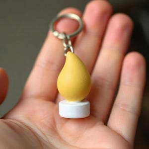 Puede incluir: Un llavero amarillo en forma de lágrima con una base blanca y un llavero plateado. El dije se sostiene en una mano, mostrando su tamaño y diseño. El color amarillo es vibrante y el dije tiene una textura suave.