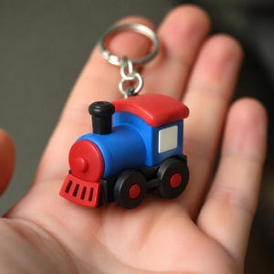 Può includere: Un portachiavi a forma di treno colorato con tetto e frontale rossi, corpo blu e ruote nere. Il portachiavi è in plastica e ha un anello in metallo argentato per attaccarlo alle chiavi. Il treno ha una piccola finestra bianca.