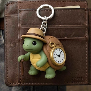 May include: A whimsical keychain featuring a turtle character wearing a hat and carrying a cane. The turtle has a clock on its shell. The keychain is attached to a silver ring and chain. The turtle is green, yellow, and brown.