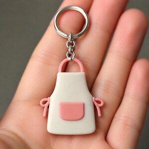 May include: A white and pink apron-shaped keychain with a silver keyring. The apron has a pink pocket and pink straps. The keychain is held in a hand.
