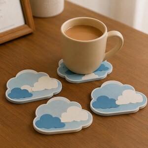 Peut inclure: Un ensemble de quatre sous-verres en forme de nuage avec des motifs superposés dans des tons bleu clair, bleu moyen et blanc. Un sous-verre soutient une tasse en céramique beige remplie d'une boisson brune, avec trois autres sous-verres disposés.