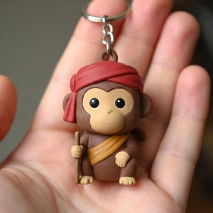 Könnte beinhalten: Ein brauner Affen-Schlüsselanhänger mit rotem Turban und gelbem Schärpe. Die Affenfigur hält einen kleinen Holzstab und hat große, schwarze Augen. Der Schlüsselanhänger ist an einem silbernen Metallring befestigt.