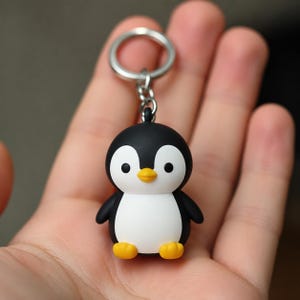 May include: A small penguin keychain featuring a smooth black and white body, a round head with black eyes, and a bright yellow beak and feet. The figure is attached to a silver-toned metal split ring by a short chain, creating a compact accessory.