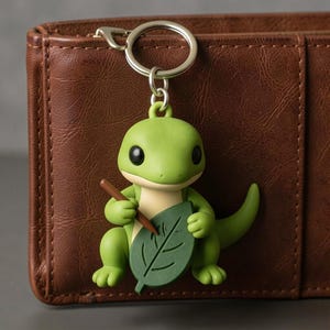 May include: A green lizard keychain with a brown pen and a green leaf. The lizard has black eyes and is holding the pen and leaf. The keychain is attached to a silver keyring. The background is a brown leather wallet.