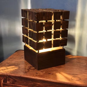 Può includere: Lampada in legno marrone scuro con design a cubo. La lampada è costruita con numerosi piccoli blocchi di legno, creando un motivo geometrico strutturato. La lampada emette una luce calda, ideale per un comodino. La lampada è alta 30 cm.