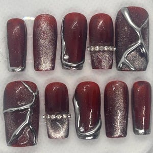 Può includere: Un set di dieci unghie finte con vari design. Le unghie sono principalmente di colore rosso scuro, con accenti di glitter argentati, linee metalliche argentate e strass. Alcune unghie sono di colore rosso pieno, altre presentano un gradiente di glitter.