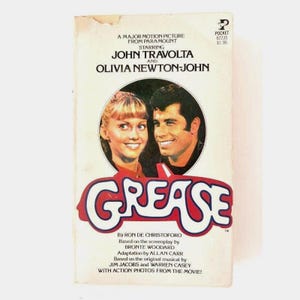 Puede incluir: Un libro de bolsillo vintage titulado Grease, que presenta un retrato circular de John Travolta y Olivia Newton-John. La portada blanca incluye el título de la película en letras rojas grandes, créditos y el logo de Pocket Books.
