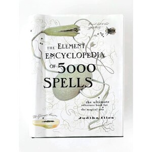 May include: A hardcover book titled The Element Encyclopedia of 5000 Spells by Judika Illes. The white cover features black text and delicate line drawings of botanical elements, including a large seed pod, vines, and a small insect illustration.
