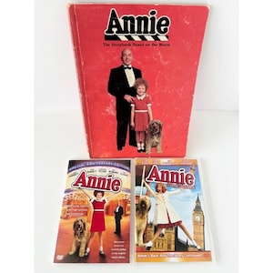 May include: A red Annie storybook featuring a man, a girl, and a dog on the cover, accompanied by two Annie DVDs. One DVD is the Special Anniversary Edition, and the other is titled Annie: A Royal Adventure, both showing movie scenes.