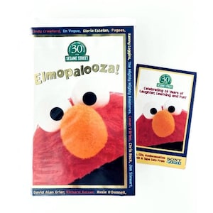 Pode incluir: Uma fita VHS Sesame Street Elmopalooza e um cartão promocional correspondente. A capa apresenta um grande rosto vermelho do Elmo com um nariz laranja brilhante e olhos brancos. O texto inclui nomes como Cindy Crawford e Sony Wonder.