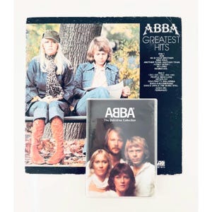 Op de afbeelding: Een ABBA Greatest Hits vinyl platenhoes met een foto van twee bandleden buiten, naast een dvd-hoesje getiteld ABBA The Definitive Collection met een portret van de vier bandleden op de voorkant.