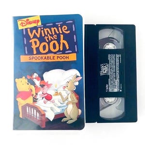 Peut inclure: Une cassette VHS Disney Winnie the Pooh Spookable Pooh avec son étui en carton bleu. La couverture montre Winnie, Tigrou, Porcinet et Coco Lapin dans une chambre. La cassette en plastique noir porte une étiquette blanche avec du texte noir.