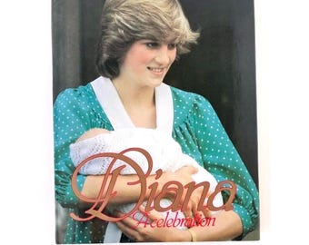 May include: A hardcover book titled Diana A Celebration featuring a photograph of a person with blonde hair wearing a green polka dot dress with a white collar, holding a baby wrapped in a white knitted blanket against a dark background.