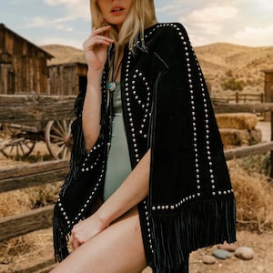 May include: Black suede cape with silver stud detailing and fringe trim. The cape has a western-inspired design with rows of studs and a flowing fringe. The cape is worn over a light green bodysuit.