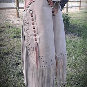 May include: Tan leather chaps with a decorative tooled belt and silver buckle. The chaps feature a fringed hem and decorative stitching along the sides. The top belt is brown with a basket weave pattern.