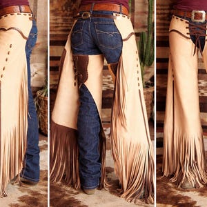 May include: Tan leather chaps with brown fringe and accents. The chaps feature decorative stitching and rivets along the sides. A brown leather belt with a silver buckle is visible at the waist. The chaps are worn over blue jeans.