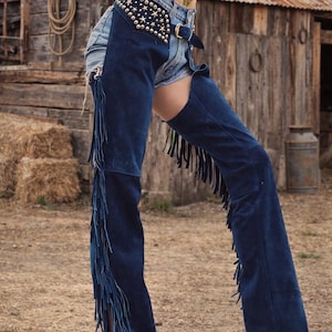 May include: Denim shorts paired with navy blue suede chaps featuring fringe detailing. The chaps are embellished with gold star-shaped studs and a belt. The image is set against a rustic, rural backdrop.
