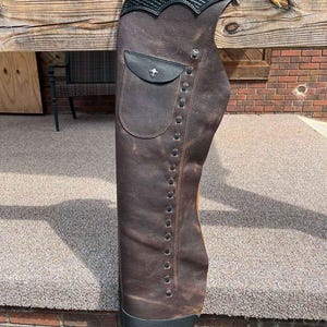 May include: Brown leather chaps with black detailing. The chaps have a pocket with a silver emblem and decorative stitching. A silver star-shaped embellishment is on the top. The chaps are designed for riding.