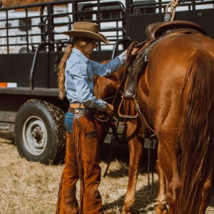 May include: A brown horse is being saddled by a person wearing a cowboy hat, blue striped shirt, and brown chaps with fringe. The saddle is brown leather. A black trailer is in the background.