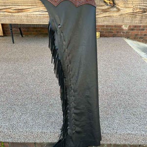 May include: Black leather chaps with brown tooled leather detailing and fringe. The chaps have a decorative silver buckle and are designed for riding. The leather is a deep black, and the fringe adds a stylish touch.