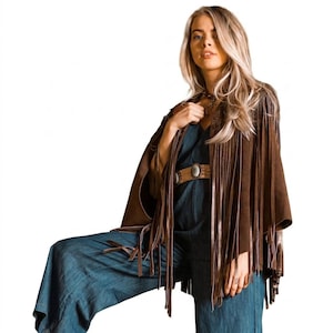 May include: A brown suede cape with long fringe detailing. The cape is worn over a denim jumpsuit and a brown belt with a large oval buckle. The cape has a bohemian, vintage style.