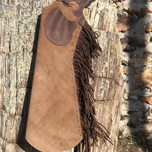 May include: A brown leather chaps with a decorative design and long fringe. The chaps have a light brown suede-like finish and are hanging against a textured wooden surface.