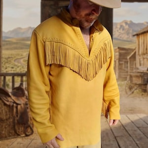 May include: A long-sleeved yellow leather shirt featuring a V-neckline and a decorative horizontal fringe panel across the chest. The garment includes braided leather detailing along the upper chest seam and additional fringe accents on the sleeves.