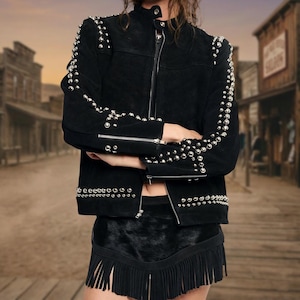 May include: A black suede jacket featuring a high collar, a front zip fastening, and silver-toned metal studs arranged along the sleeves and hem. The jacket is paired with a black skirt that has a textured upper section and a long fringe hemline.