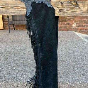 Könnte beinhalten: Schwarze Wildleder-Chaps mit einem dekorativen Blumendesign am oberen Teil. Die Chaps haben lange Fransen an den Seiten und einen Gürtel mit einer Schnalle. Die Chaps hängen an einem Holzbalken.