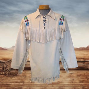 May include: A cream-colored suede shirt featuring long fringe detailing along the chest, sleeves, and hem. The garment includes a lace-up front closure and colorful geometric beadwork accents on the shoulders, providing a textured appearance.