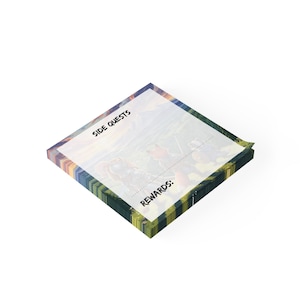 May include: A square notepad with a colorful border and the words "SIDE QUESTS" and "REWARDS" printed on the top. The notepad features an illustration of a fantasy scene. The notepad is on a white background.