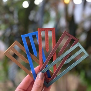 May include: A hand holds a fan of colorful, geometric paper clips. The clips are in shades of gold, blue, red, brown, and teal. Each clip has a rectangular frame with a central, pointed extension.