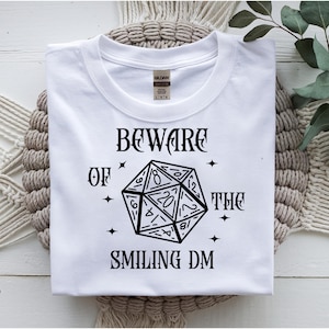 May include: White t-shirt with the text "BEWARE OF THE SMILING DM" and a black illustration of a twenty-sided die. The shirt is made of a soft material and is suitable for casual wear. The shirt is folded on a beige surface.