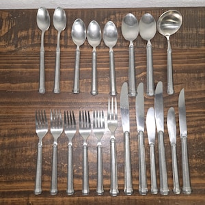 May include: A collection of vintage silverware, including spoons, forks, knives, and a ladle, arranged on a wooden surface. The silverware has a matte silver finish and a simple, elegant design. The forks have four tines.