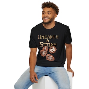 May include: Black t-shirt with the text "Unearth A Story" in a decorative font. The shirt features illustrations of dinosaur fossils in brown and tan colors. The person is wearing blue jeans.