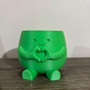 May include: A bright green planter with a smiling face, arms forming a heart shape, and small feet. The planter is made of a smooth material and sits on a wooden surface. The design is whimsical and cheerful.