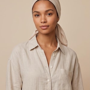 Puede incluir: Un pañuelo beige atado en la parte posterior de la cabeza, combinado con una camisa beige de manga larga con botones, cuello clásico y un bolsillo en el pecho. La tela tanto del pañuelo como de la camisa es de un tono sólido y neutro.