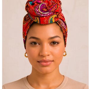 Puede incluir: Un pañuelo para la cabeza con un vibrante estampado de cachemira en tonos rojo, morado, amarillo y turquesa. La tela está atada en un nudo estructurado en la parte superior de la cabeza, cubriendo el cabello por completo. Se ven pequeños aros dorados.