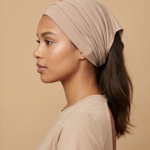 May include: A beige head wrap, with a gathered design. The head wrap is made of a soft, stretchy material. The person is wearing a beige t-shirt. The background is a solid beige colour.