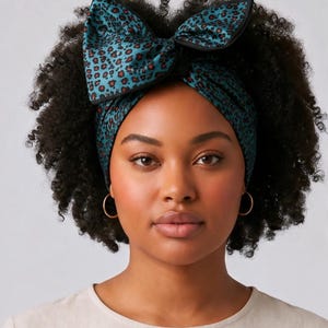 Peut inclure: Un bandeau bleu sarcelle avec un motif léopard noir et un grand nœud structuré sur le dessus. Le tissu présente une finition lisse avec un liseré noir sur les bords du nœud, créant un contraste marqué avec le fond bleu sarcelle.