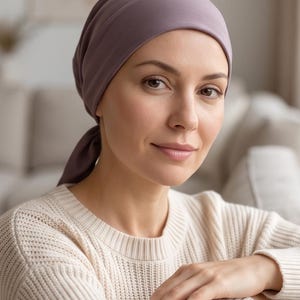 Könnte beinhalten: Ein einfarbiges, malvenfarbenes Kopftuch, das am Hinterkopf gebunden ist, kombiniert mit einem cremefarbenen Langarmpullover mit strukturiertem Strickmuster. Das Kopftuch besteht aus einem glatten, matten Stoff, der das Haar vollständig bedeckt.