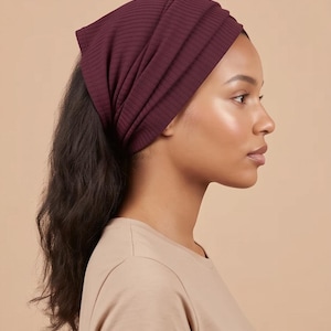 May include: A deep burgundy head wrap made of a ribbed fabric. The head wrap features a series of horizontal folds and gathers that create a textured, layered appearance across the crown, covering the top of the head while leaving hair exposed.