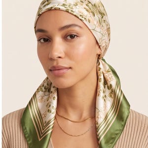 Puede incluir: Un pañuelo cuadrado con base color crema y un estampado botánico de hojas verdes y flores de color naranja claro. El pañuelo tiene un borde ancho de color verde oscuro con finas rayas blancas en los extremos, atado a la cabeza.