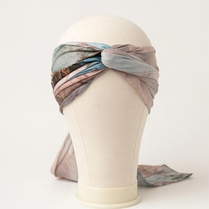 May include: A patterned fabric headband with a twisted knot design. The headband features a watercolour-like pattern in shades of blue, grey, and brown. The headband is displayed on a mannequin head against a white background.